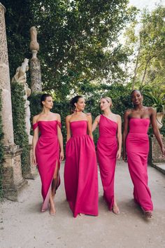 three women in bright pink dresses walk down a path near some trees and stone pillars