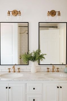 two sinks in a bathroom with mirrors and lights on the wall above them, along with a vase filled with flowers