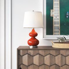 an orange lamp sitting on top of a wooden cabinet next to a painting and basket