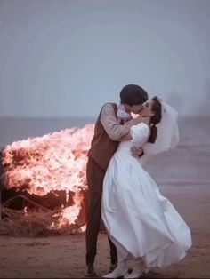 a bride and groom kissing in front of a fire on the beach with their arms around each other