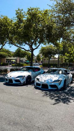 two white sports cars parked next to each other in a parking lot with trees behind them