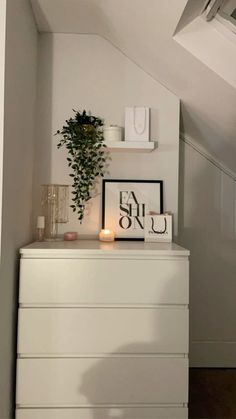 a white dresser topped with a plant and candles next to a wall mounted art piece