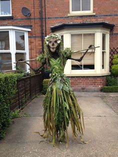 a person dressed up as a skeleton with plants on their head and arms, standing in front of a brick building