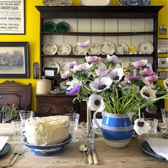 a table topped with a blue vase filled with purple and white flowers next to a cake