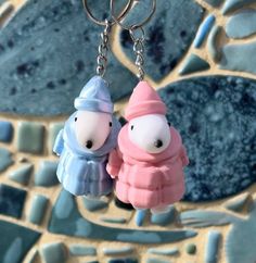 two little toy figures are hanging from a key chain on a mosaic tile background, one is wearing a pink hat and the other is wearing a blue coat