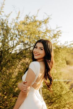 a woman in a white dress is posing for a photo with trees and bushes behind her