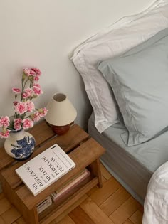 a bed with white sheets and pink flowers in a vase next to a book on a wooden table