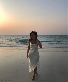 a woman in a white dress walking on the beach at sunset with her cell phone to her ear