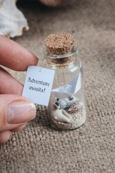 a person holding a tiny bottle filled with sand and sea shells next to a tag that says adventure awaits