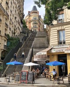 people are sitting at tables on the sidewalk in front of some buildings with blue umbrellas