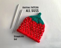 a knitted strawberry hat next to two knitting needles on top of a white surface