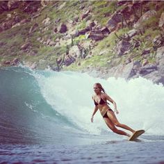 a woman riding a surfboard on top of a wave in front of a mountain