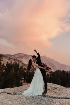 a bride and groom standing on top of a mountain with their arms in the air