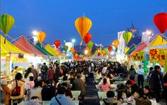 many people are sitting at tables in an outdoor market with colorful lanterns hanging from the ceiling