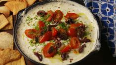 a bowl of dip with tomatoes and olives in it next to chips on the table