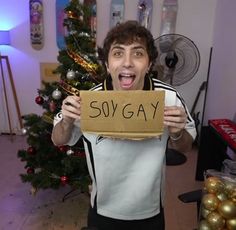 a man holding up a cardboard sign with the words so gay on it in front of a christmas tree