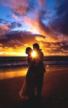 a man and woman standing on top of a beach next to the ocean at sunset