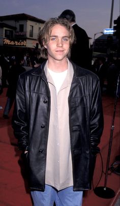 a man in a black leather jacket and jeans standing on a red carpet with his hands in his pockets