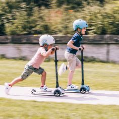 two young boys are riding scooters on the road in front of some trees