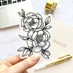 a person holding up a flower sticker next to a laptop and pen on a desk