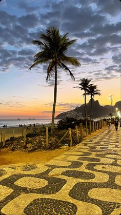 people are walking on the sidewalk next to the ocean at sunset with palm trees in the foreground