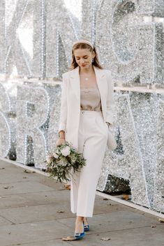 a woman in a white suit and blue shoes is walking down the street with her bouquet