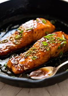 two pieces of meat in a skillet with spoons on the side and sesame seeds sprinkled on top