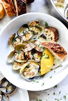 clams in a white bowl with bread on the side and another dish next to it