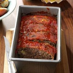 a meatloaf in a pan on a wooden table with other food and utensils