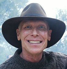 a man wearing a black hat and sweater smiling at the camera with trees in the background