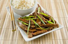 a white plate topped with meat and rice next to chopsticks on a bamboo mat