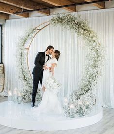 a bride and groom kissing in front of a white backdrop with greenery on it