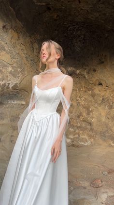 a woman in a white wedding dress standing next to a stone wall with her hands on her hips