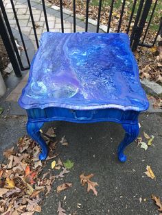 a blue table sitting on top of a sidewalk next to a black iron fence and leafy ground