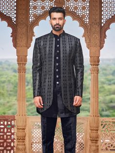 a man standing in front of a wooden structure wearing a black and white suit with an intricate