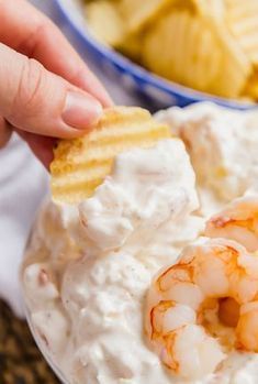 a person dipping shrimp into a bowl of dip with crackers on the side and chips