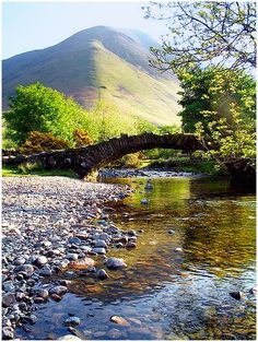 a river running through a lush green forest next to a stone covered bridge with a mountain in the background