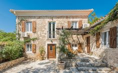 an old stone house with wooden shutters on the front and side doors, surrounded by greenery