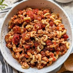 a bowl filled with pasta and meat on top of a striped table cloth next to a wooden spoon