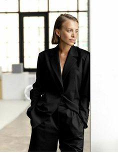 a woman in a black suit standing next to a wall and looking at the camera