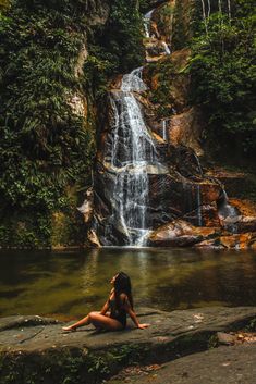 a woman sitting on the edge of a river in front of a waterfall with green foliage