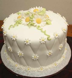 a white cake with daisies on it sitting on top of a wooden table next to a wall