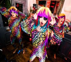 three people dressed in brightly colored clothing and one person with bright hair is dancing on the floor