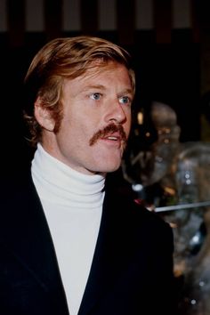 a man with a moustache wearing a black suit and white turtle neck sweater