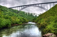 a bridge over a river surrounded by lush green trees and mountains under a cloudy sky