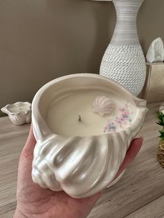 a hand holding a white candle in a shell shaped bowl with shells on the inside