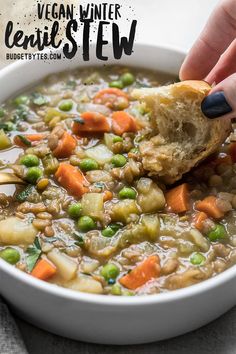 a person dipping a piece of bread into a bowl of vegetable soup with peas and carrots