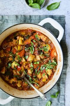 a large pot filled with stew and vegetables on top of a blue cloth next to a spoon