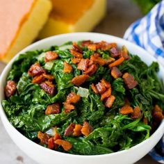 a white bowl filled with greens and bacon on top of a table next to bread