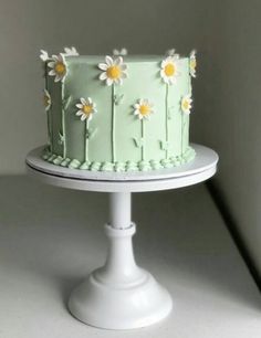 a green cake with daisies on it sitting on top of a white pedestal next to a wall